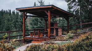 A wooden gazebo with a brick fireplace, surrounded by a wooden fence and dense forest.
