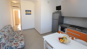 Eine kleine Küche mit Kühlschrank, Herd und Mikrowelle. Ein Esstisch mit Obstschale und ein Sofa befinden sich im Wohnbereich.