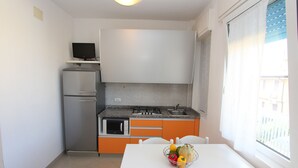 A compact kitchen with a refrigerator, microwave, stove, and sink. A table with a fruit bowl is in front of a window. - Flat a few steps from the beach (Bibione)