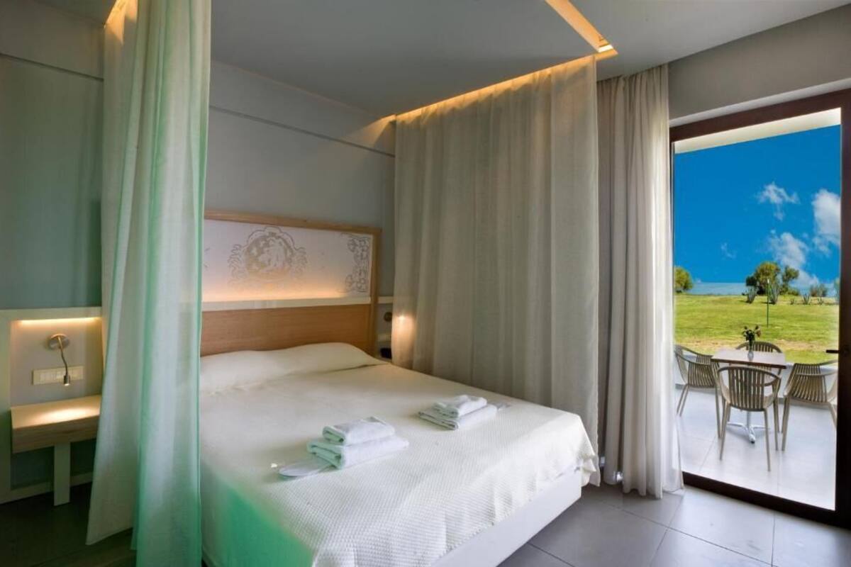 A hotel room with a large bed, a bedside table, a view of a grassy area through a glass door, and a table with chairs outside.