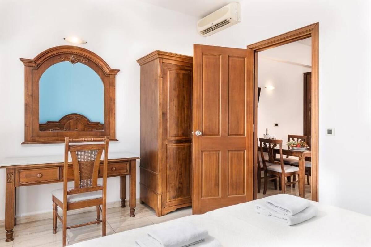 A bedroom with a wooden wardrobe, a desk with a mirror, and a dining area visible through an open door.