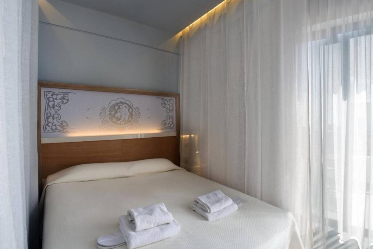 A neatly made bed with white linens and towels, a headboard with a decorative panel, and sheer curtains allowing natural light.