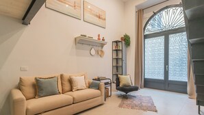 Sala de estar com um sofá bege, uma poltrona preta, uma mesinha com um livro e velas, uma estante com objetos decorativos e uma janela grande com vista.