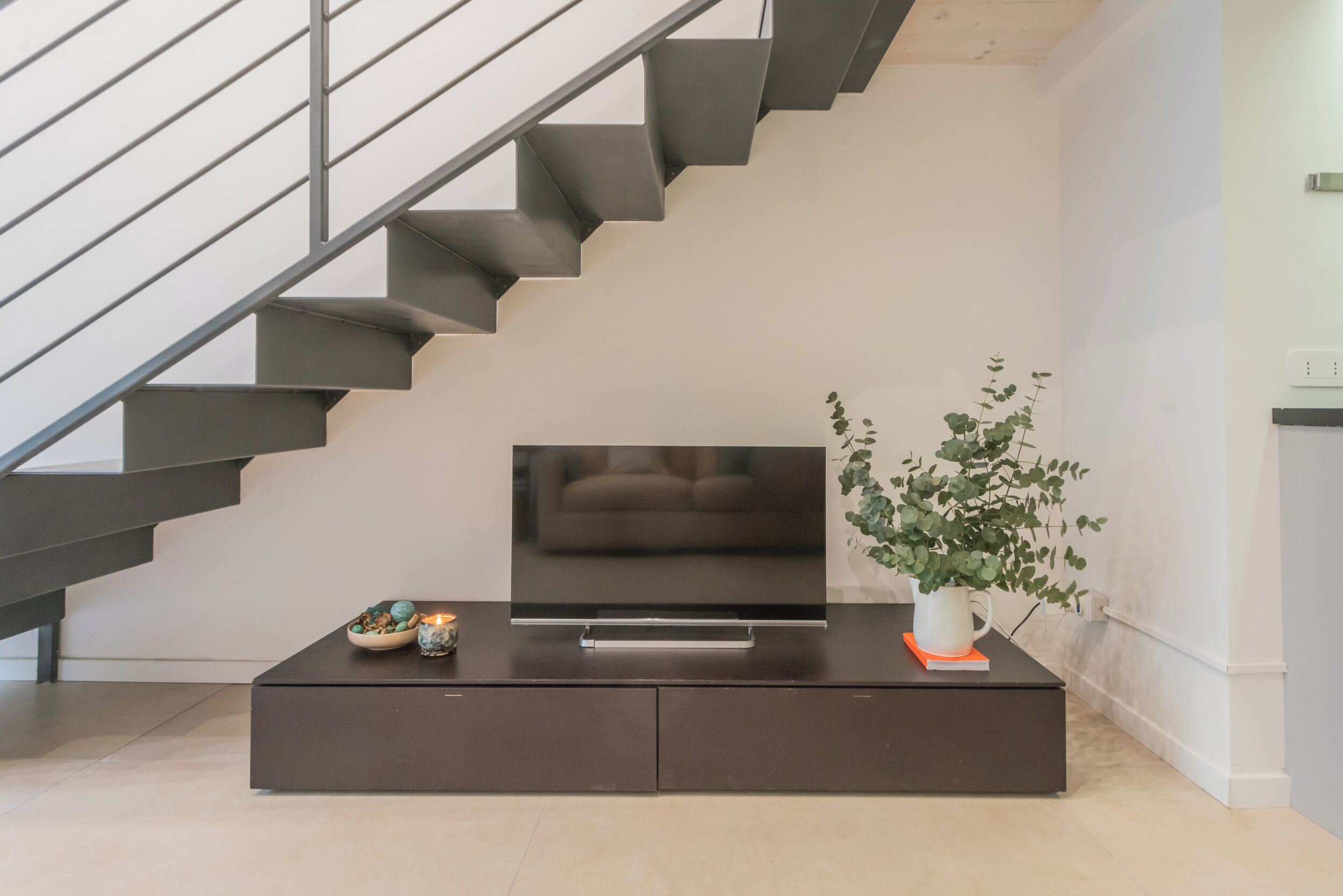 A modern living room with a large flat-screen TV on a dark wooden console, a staircase with dark railings, and a potted plant on the console.
