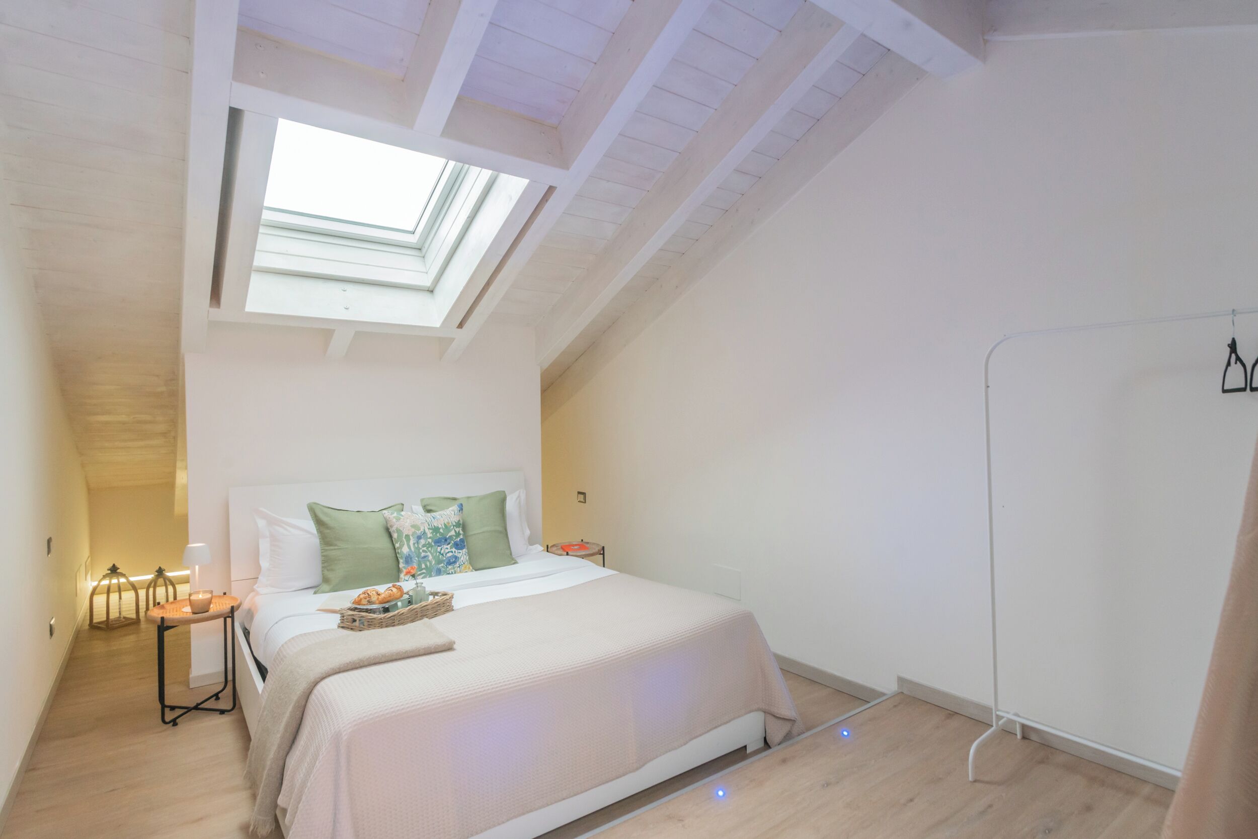 A bedroom with a skylight, a bed with pillows, a bedside table with a lamp, and a small table with a basket.