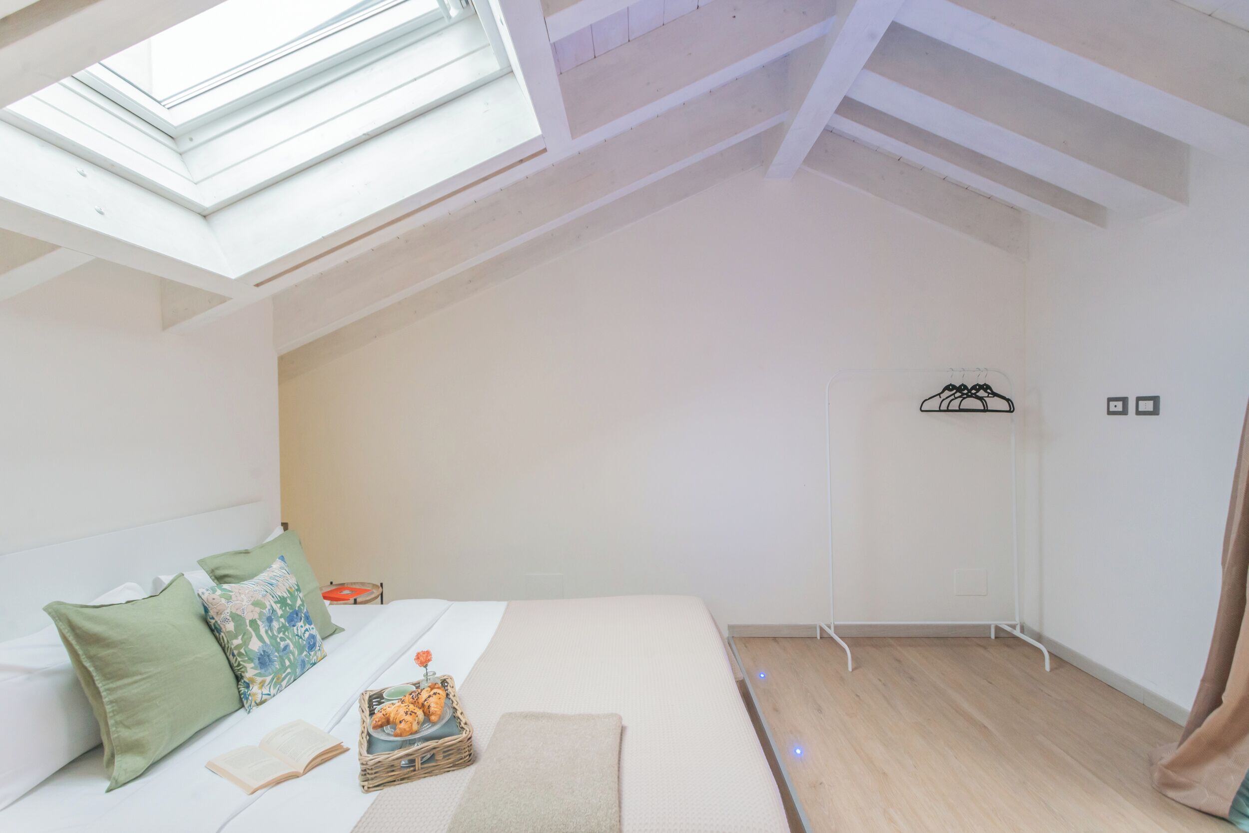 A bedroom with a skylight, a bed with pillows, a bedside table with a book and a basket, and a wooden floor.
