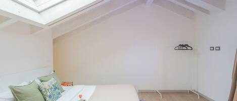 A bedroom with a skylight, a bed with pillows, a bedside table with a book and a basket, and a wooden floor.