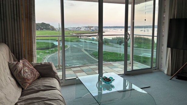 A modern living room with a large sliding glass door leading to a view of a bridge and water. - Mudeford, Dorset. Spectacular Seafront Views. Perfect Family Getaway. Driveway. (Christchurch)