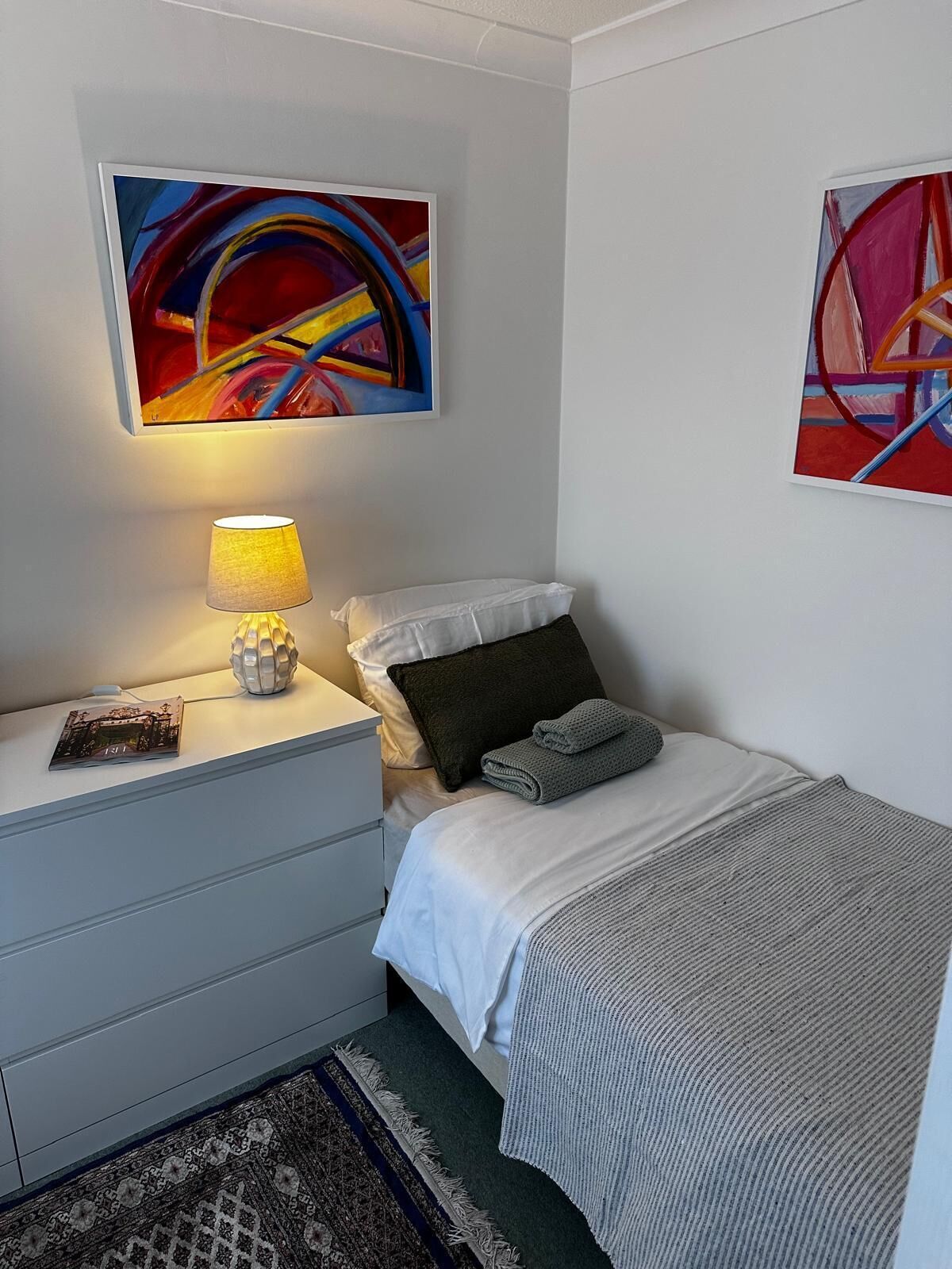 A bedroom with a bed, a nightstand, a lamp, and two abstract paintings on the walls.
