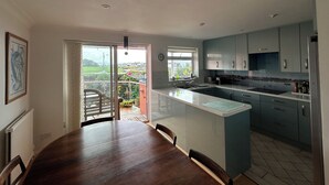 A modern kitchen with a large island, a dining area with a wooden table, and a view of a garden through a sliding door. - Mudeford, Dorset. Spectacular Seafront Views. Perfect Family Getaway. Driveway. (Christchurch)