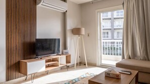 Een moderne woonkamer met een flatscreen televisie, een houten paneelwand, een staande lamp en een groot raam met uitzicht op een gebouw.