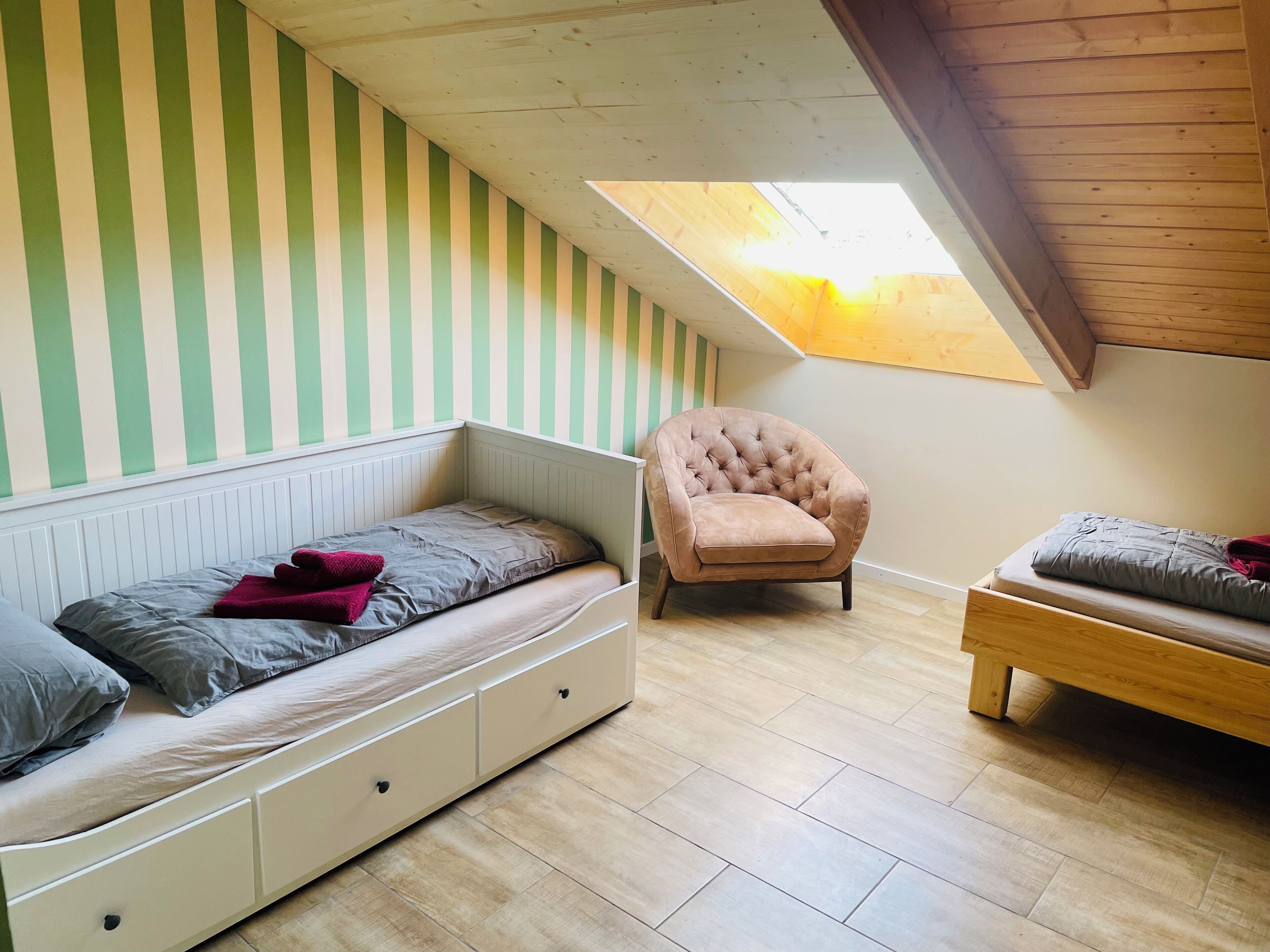 A small, cozy room with a sloped ceiling, a single bed with a white frame and storage drawers, a cushioned armchair, and a second bed with a wooden frame.