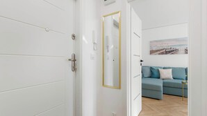 A modern room with a wooden floor, a white door with a gold frame, a blue sofa with cushions, and a painting of a beach scene. - Cozy Apartment with Bedroom | Close to Park and Beach (Gdańsk)