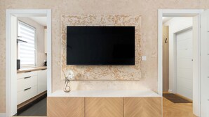 A modern kitchen with a large flat-screen TV mounted on the wall, wooden cabinets, and a white countertop. - Cozy Apartment with Bedroom | Close to Park and Beach (Gdańsk)