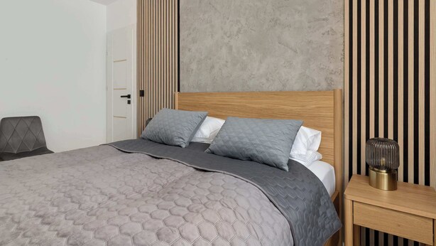 A modern bedroom with a large bed, a bedside table, and a wall with vertical wooden slats. - Cozy Apartment with Bedroom | Close to Park and Beach (Gdańsk)