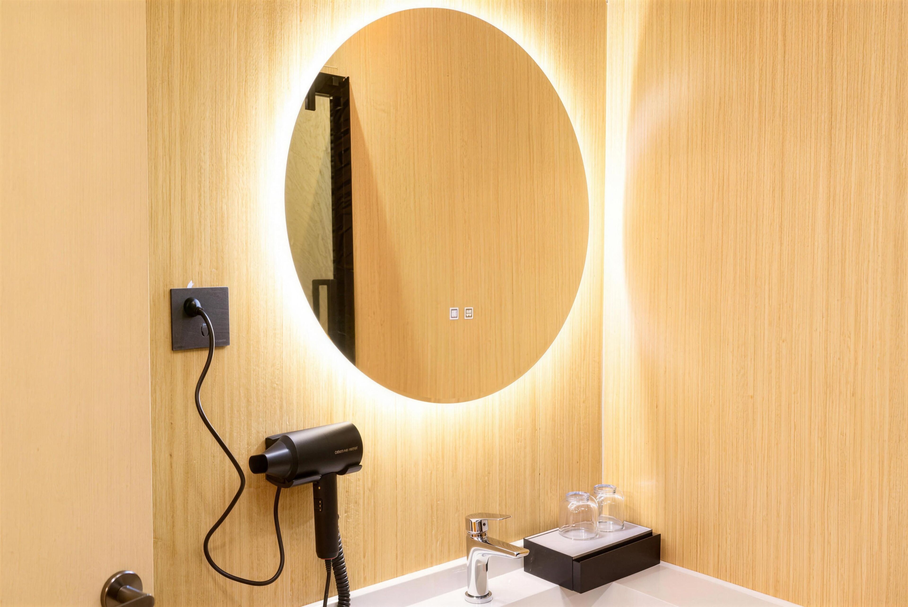Un baño con pared de madera, un espejo redondo, un secador de pelo y un lavamanos con recipientes de vidrio.