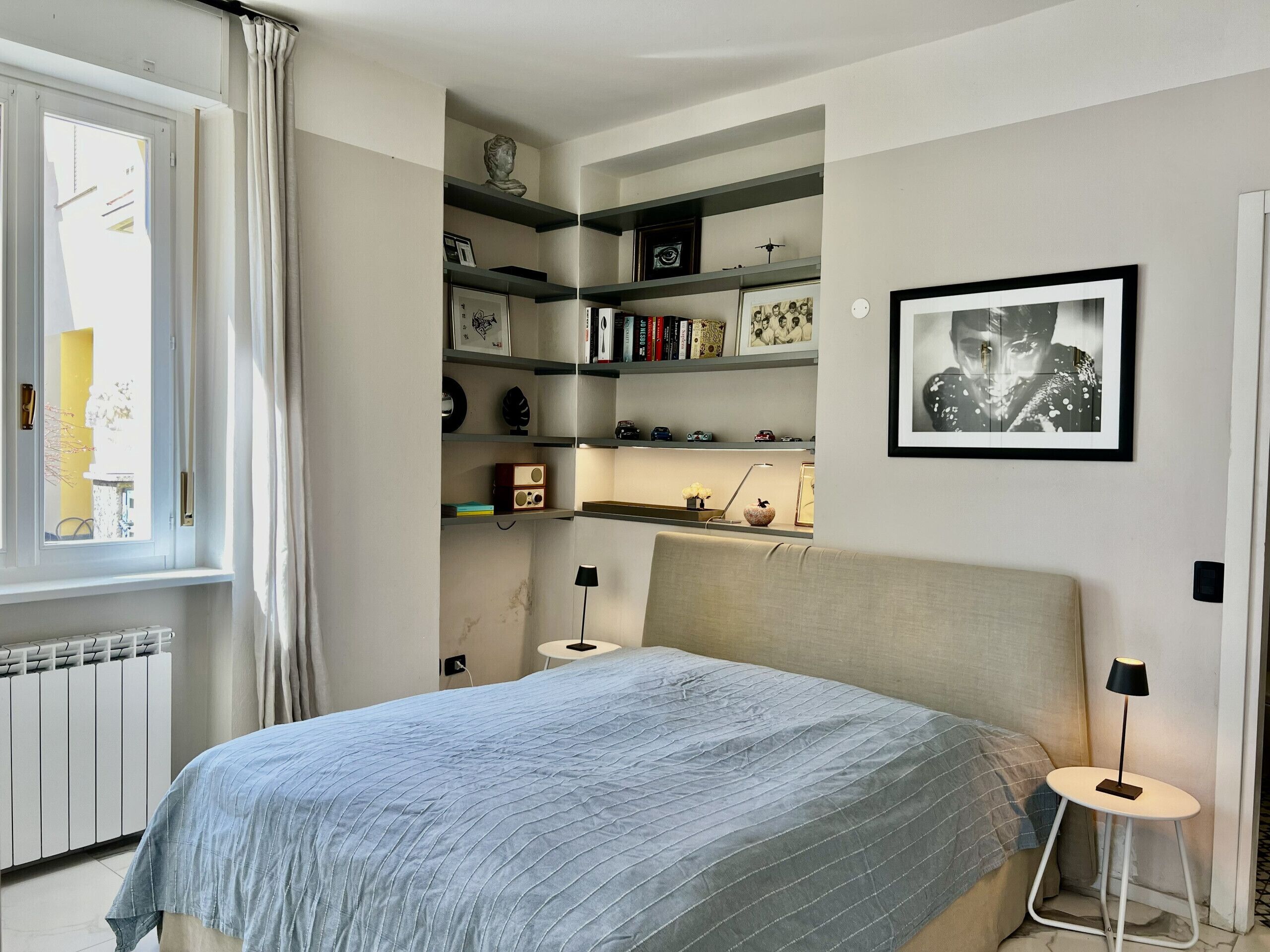 A bedroom with a bed, a bedside table, a lamp, a radiator, and a window with curtains.