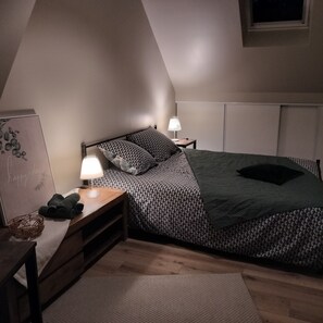 Ett sovrum med en säng, två sängbord, ett litet bord, en lampa, en hylla som är monterad på väggen och ett fönster.