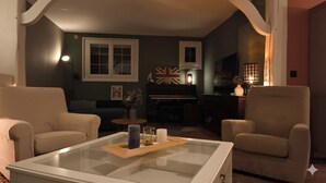 Ein Wohnzimmer mit zwei beigefarbenen Sesseln, einem Glastisch mit Kerzen, einem Klavier und einer Lampe.