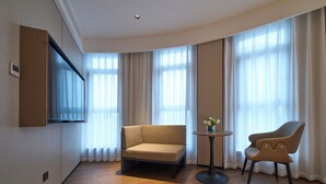 Een moderne hotelkamer met een bank, een stoel, een klein tafeltje met bloemen en een flatscreen televisie aan de muur.