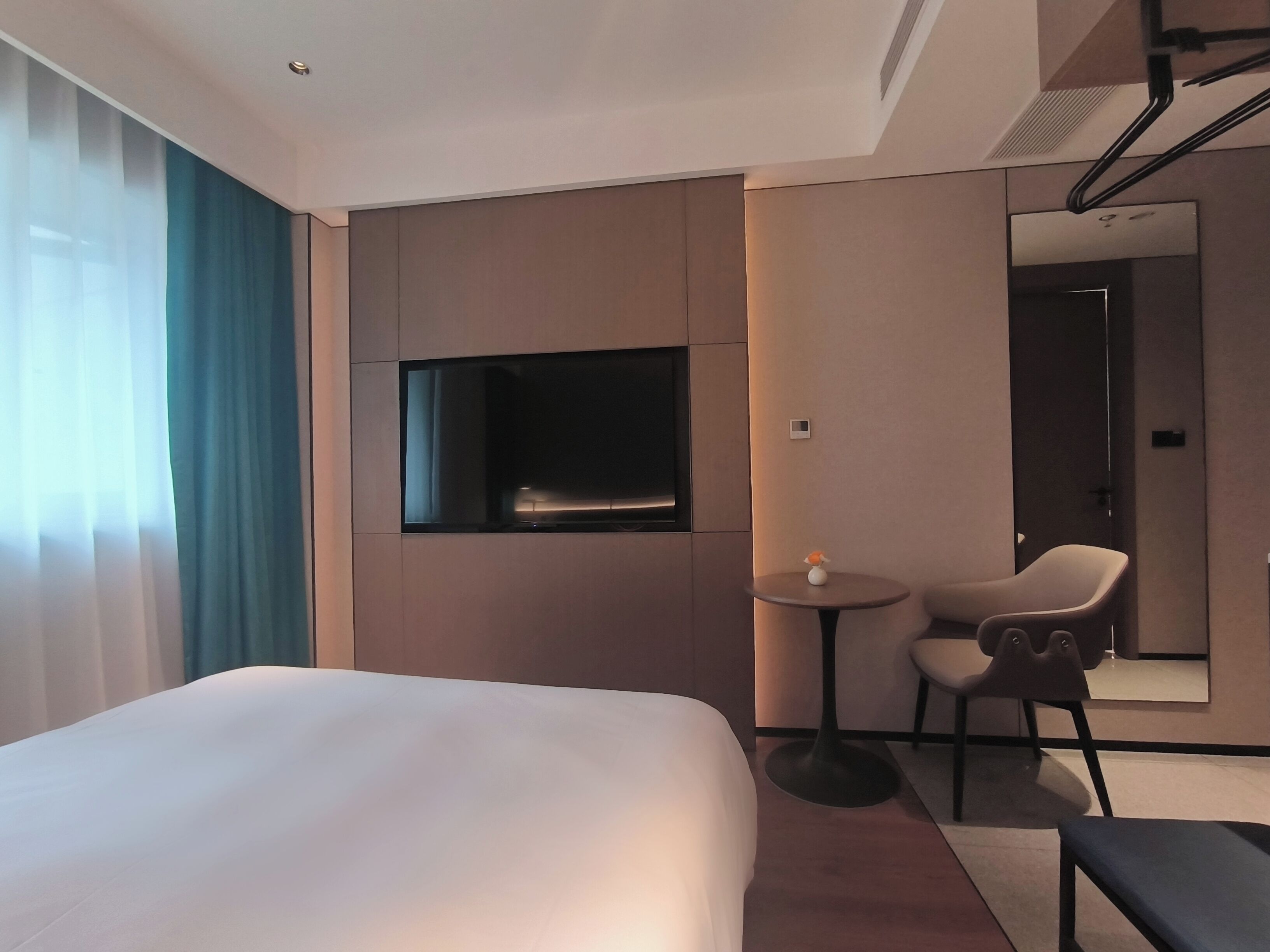 A hotel room with a bed, a TV mounted on the wall, a small table with a chair, and a mirror.