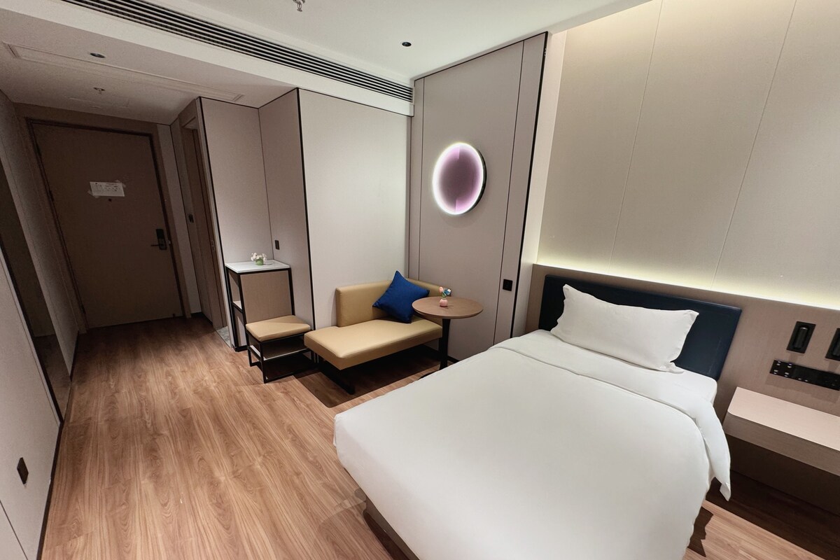 A hotel room with a large bed, a small seating area with a table, a wall-mounted light, and a door.