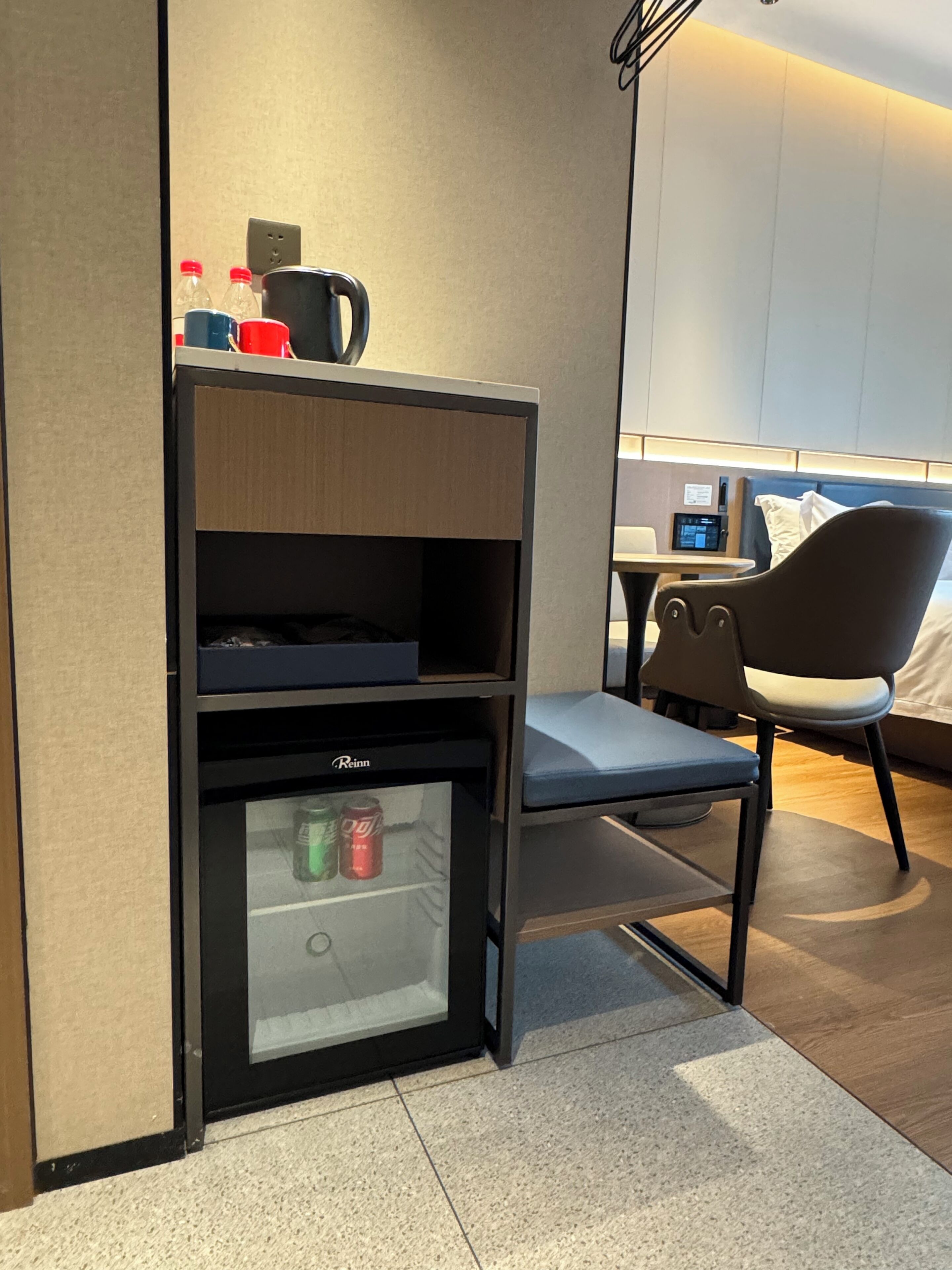 Habitació d'hotel compacte amb minibar, nevera petita, una cadira i un llit amb capçalera.
