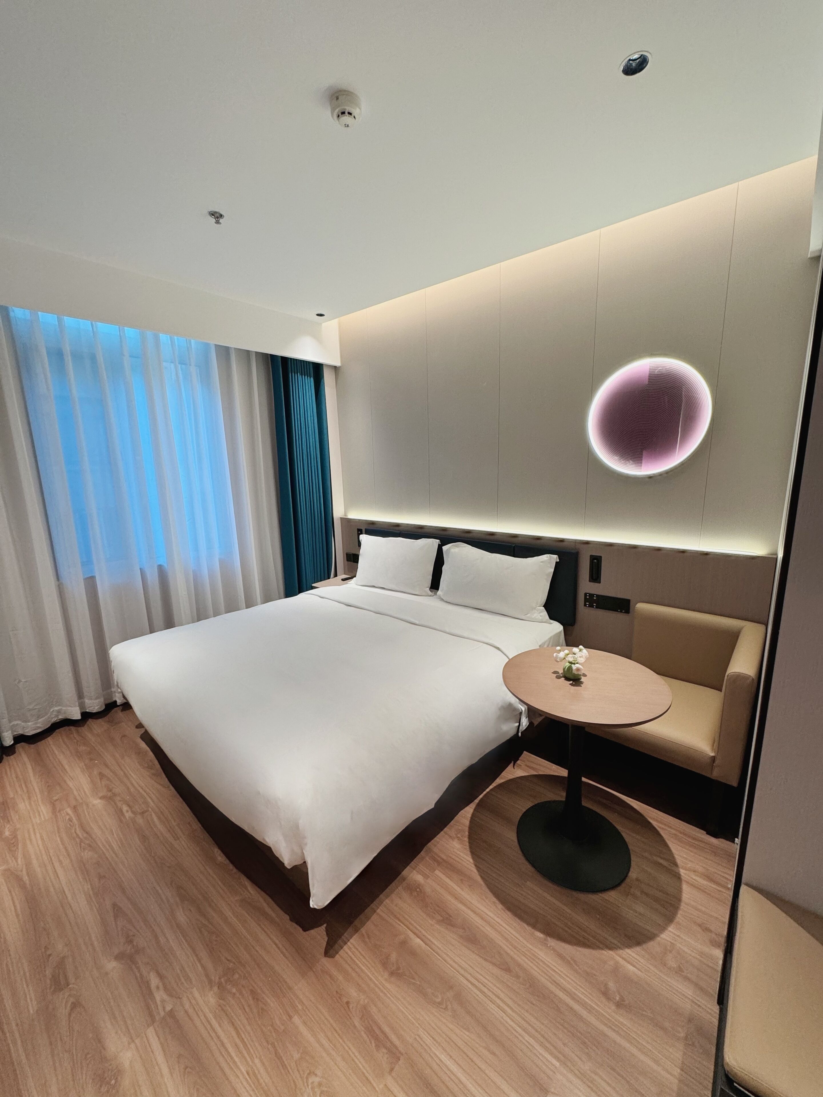 A modern hotel room with a large bed, a round table, a sofa, and a ceiling-mounted light fixture.