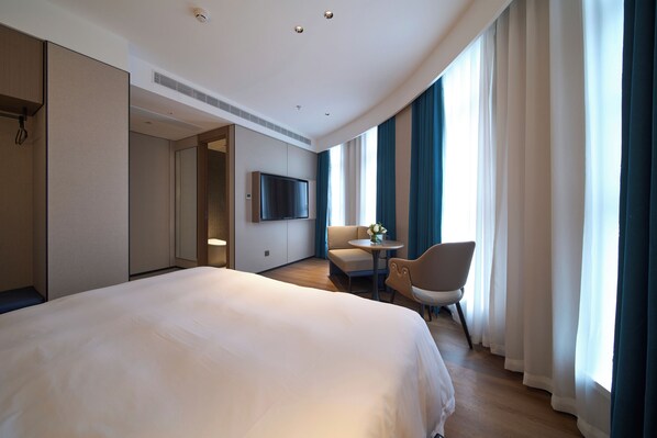 Ein Hotelzimmer mit einem großen Bett, einem Fernseher, einem kleinen Tisch mit Stuhl und Blick durch Vorhänge nach draußen.