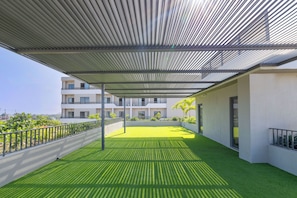 A covered outdoor area with artificial grass, a metal railing, and a building in the background. - The Bantree (Accra)