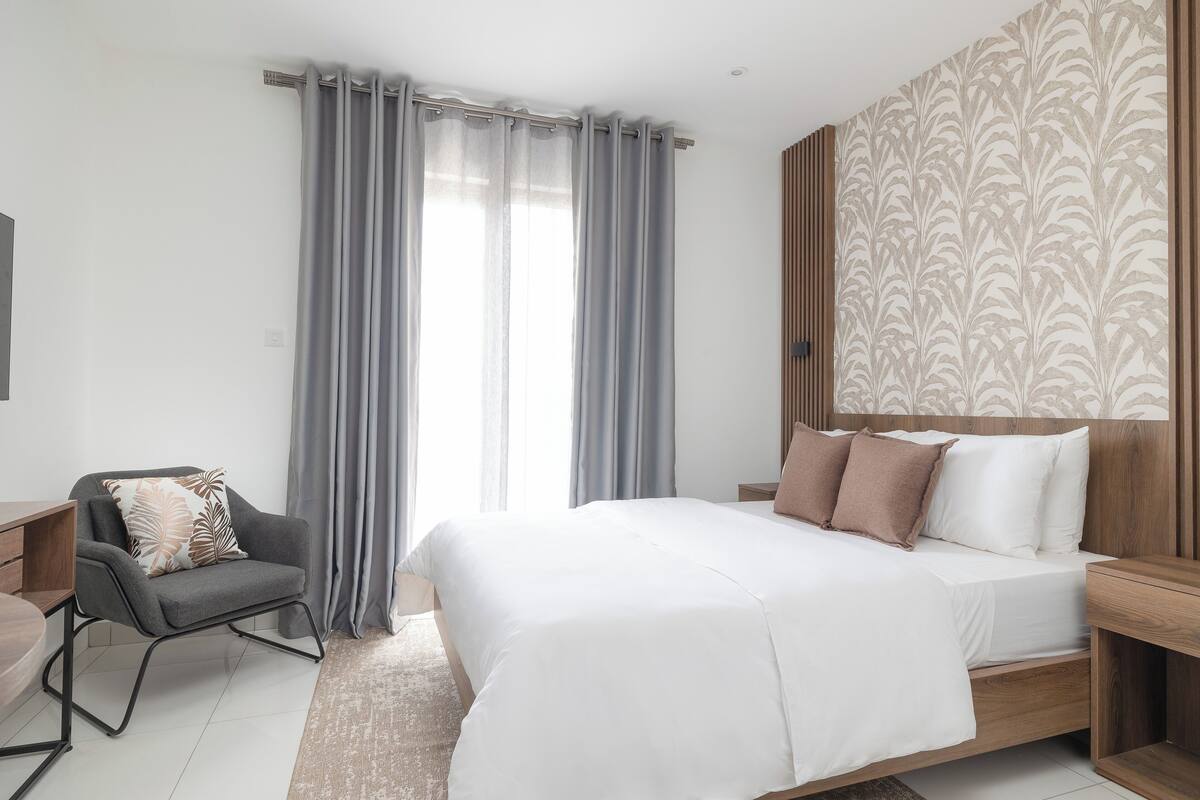 A modern hotel room with a large bed, a grey armchair, and a wooden headboard.
