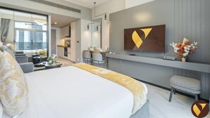Moderni hotellihuone, jossa on suuri sänky, seinälle asennettu televisio, pieni oleskelualue ja näkymä kaupunkiin.