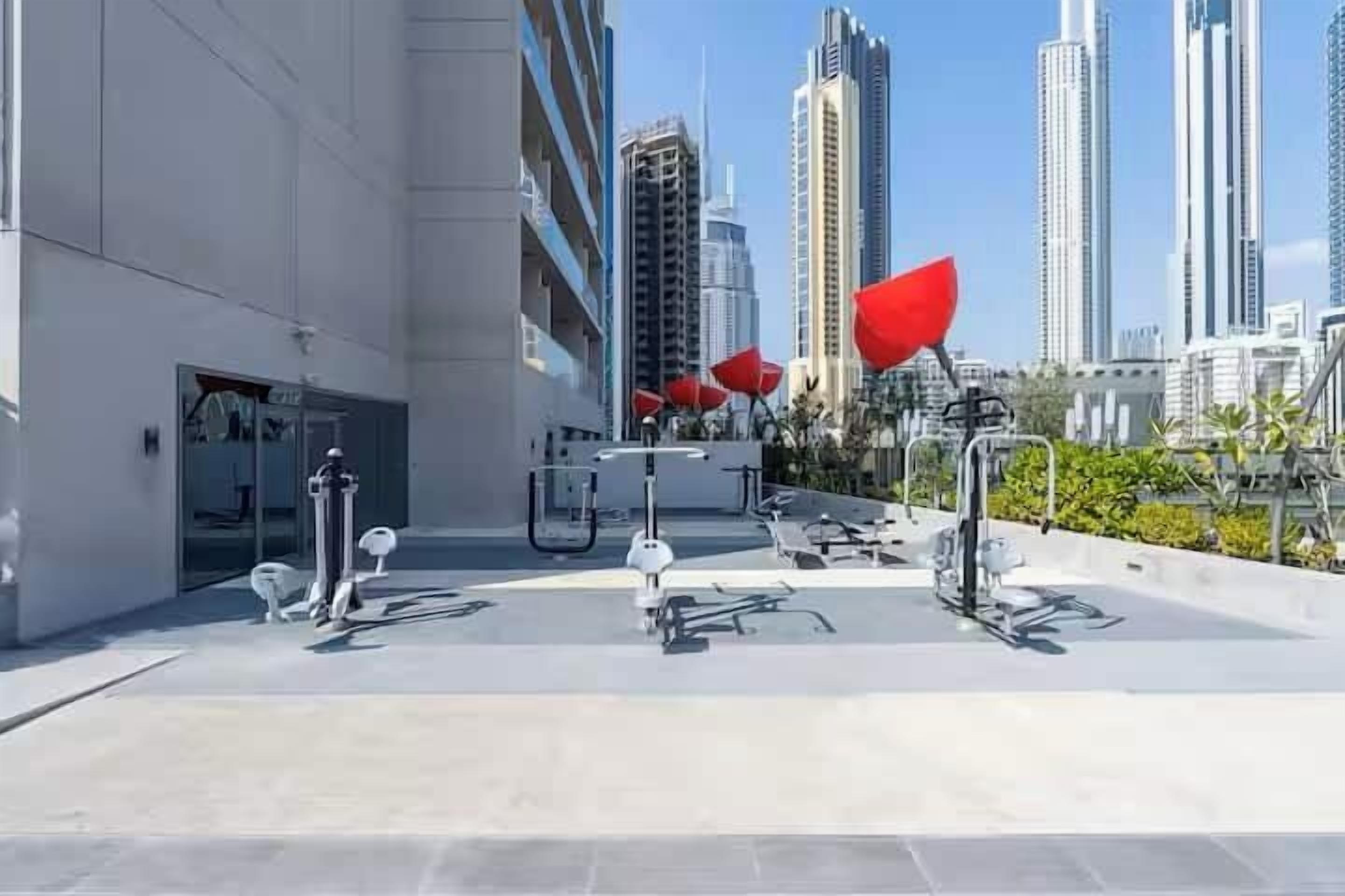 Un espacio exterior moderno con equipamiento de ejercicio, un acceso de edificio de vidrio y rascacielos al fondo.