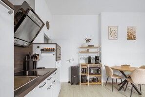 Eine kompakte Küche mit Backofen, Kaffeemaschine und kleinem Kühlschrank. Es gibt eine Essecke mit einem Holztisch und Stühlen.