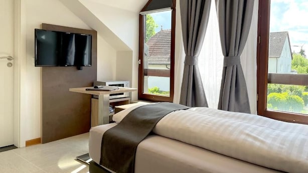 A bedroom with a bed, a TV mounted on the wall, a desk with a computer, and large windows with curtains. - Hana Aparthotel (Barsbuttel)