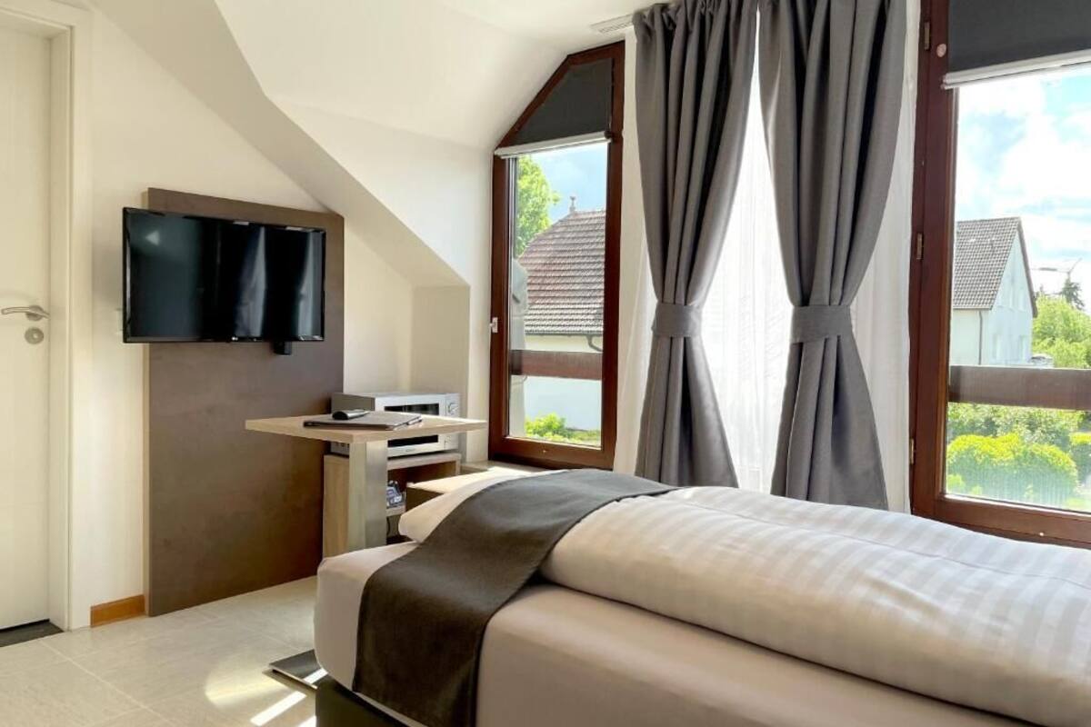 Una camera da letto con un letto, una TV a parete, una scrivania con un computer e ampie finestre con tende.
