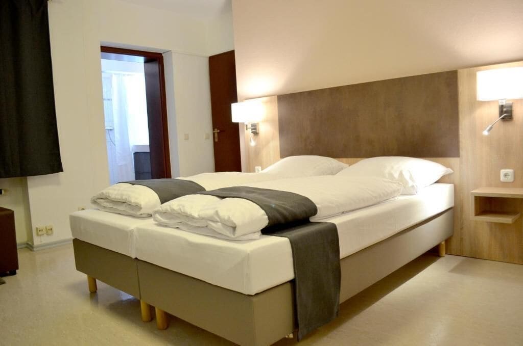 Cama doble con ropa de cama blanca y gris, una mesita de noche con lámpara y un cabecero de madera.