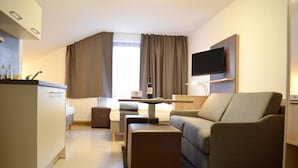 A modern hotel room with a kitchenette, a sofa, a coffee table, and a flat-screen TV mounted on the wall. - Hana Aparthotel (Barsbuttel)