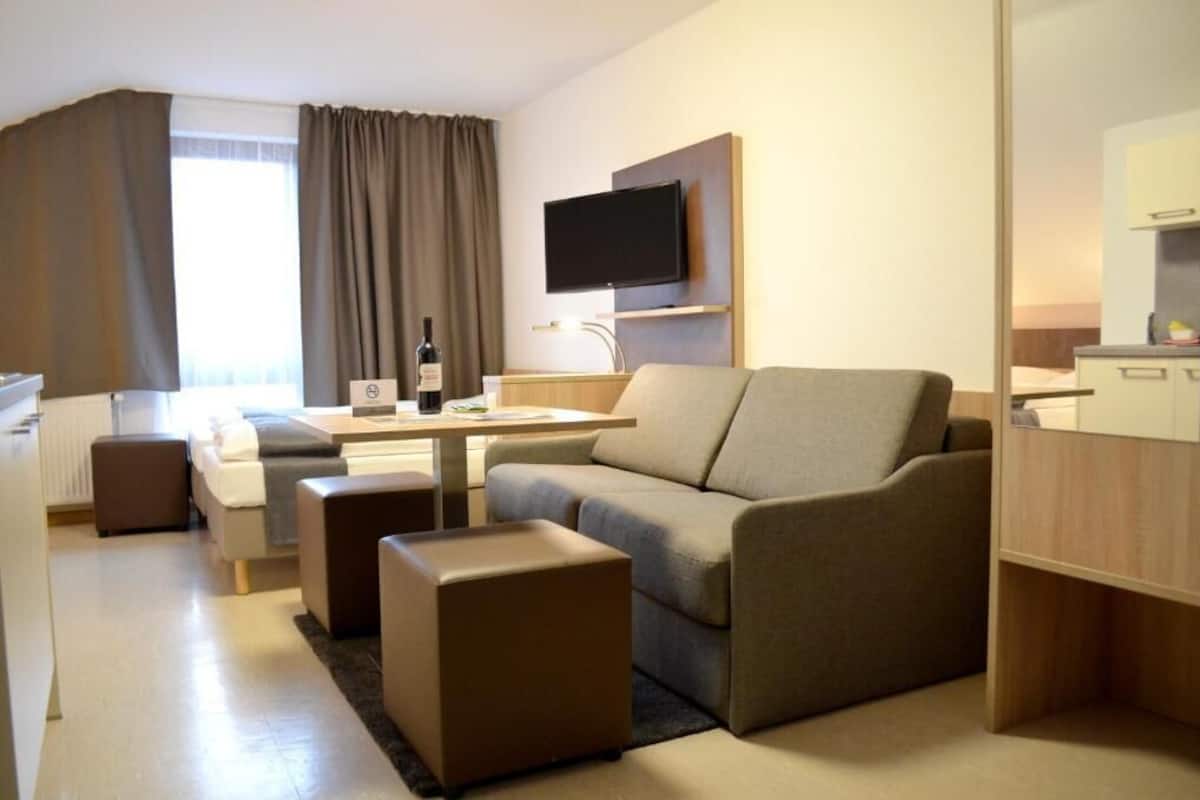 Una moderna camera d'albergo con divano, un piccolo tavolo, una TV a schermo piatto fissata al muro, un minibar e una finestra con tende.