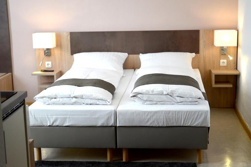 A double bed with white linens and a dark headboard, flanked by bedside tables with lamps, in a modern hotel room.