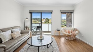 A modern living room with a sofa, a round coffee table, and a wicker chair. There are two large windows offering a view of the outdoors. - Modern Townhouse in Wright with Mt Stromlo Views (Wright)