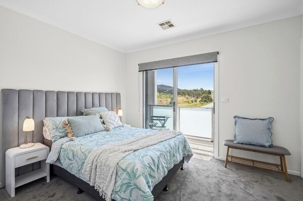 A bedroom with a large bed, a nightstand, a bench, and a view of the outdoors.