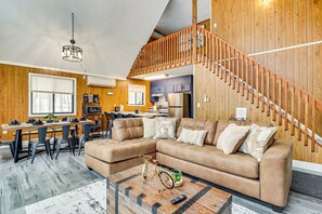 A cozy living room with a large beige sectional sofa, a wooden staircase, and a kitchen in the background.