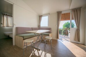 A compact living space with a convertible sofa, a small table, and two stools. A window provides natural light and a view of the outdoors. - Mobile Home for Families at Lake Garda (Moniga del Garda)