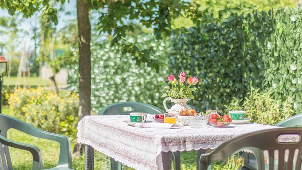 Una mesa dispuesta al aire libre con mantel floreado, una jarra de flores y diversos alimentos para el desayuno.