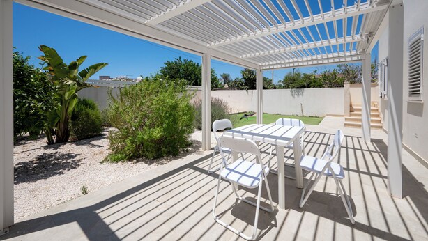 Un patio techado con una mesa y sillas, rodeado de follaje y un cielo azul despejado.