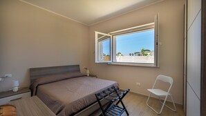 Un dormitorio con cama, una silla, un escritorio y una ventana con vistas a edificios y palmeras.