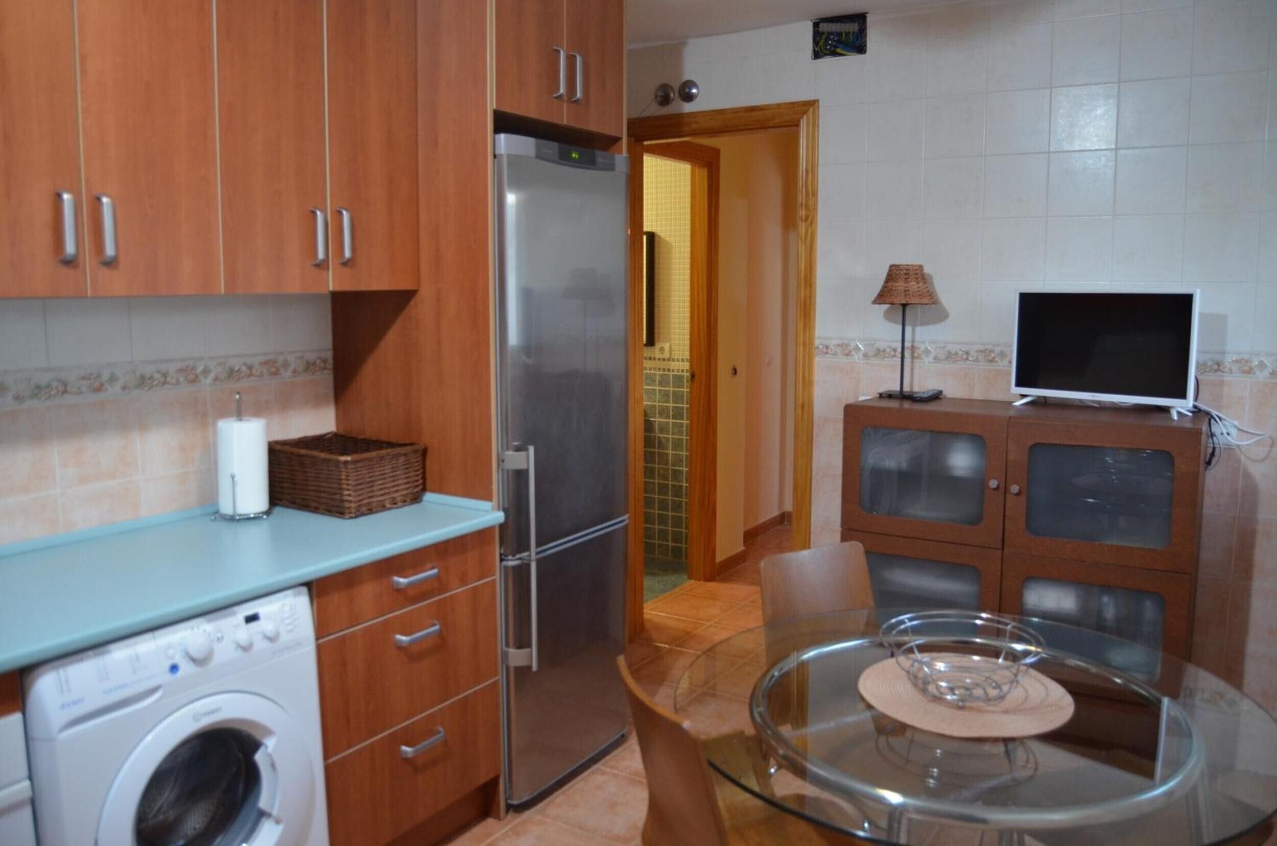 Una cocina compacta con armarios de madera, un refrigerador de acero inoxidable, lavadora, una mesa de comedor con sillas y un televisor sobre un mueble.