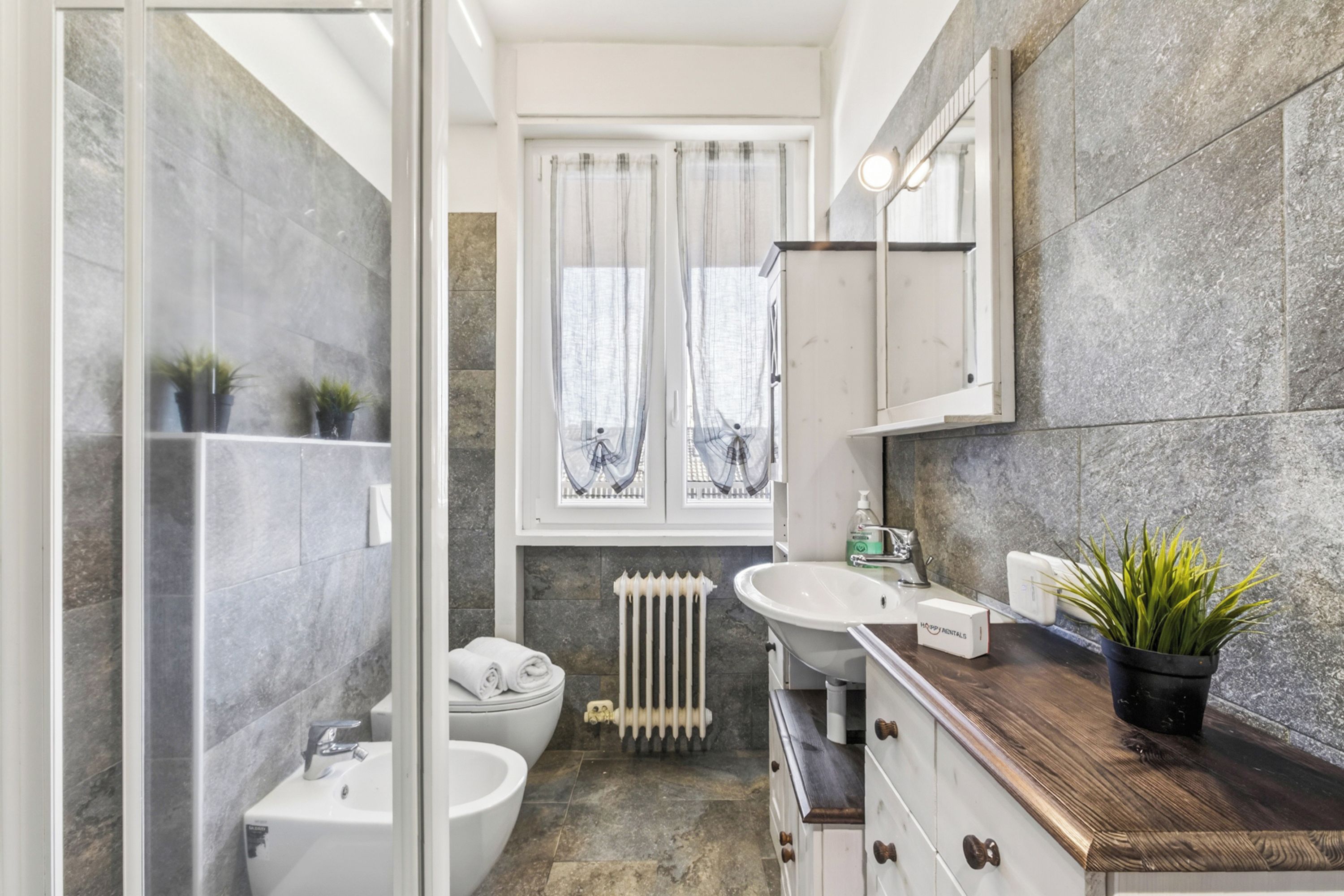 A bathroom with a glass shower enclosure, a white sink, a toilet, a radiator, and a wooden vanity with a mirror.