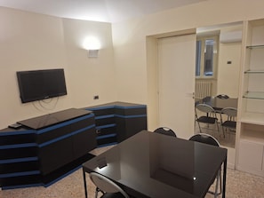 Ein Zimmer mit einem an der Wand befestigten Flachbildfernseher, einem schwarzen Tisch mit Stuhl und einem kleinen Schreibtisch mit Schubladen. Eine Tür führt in einen weiteren Raum mit Fenster und Heizkörper.